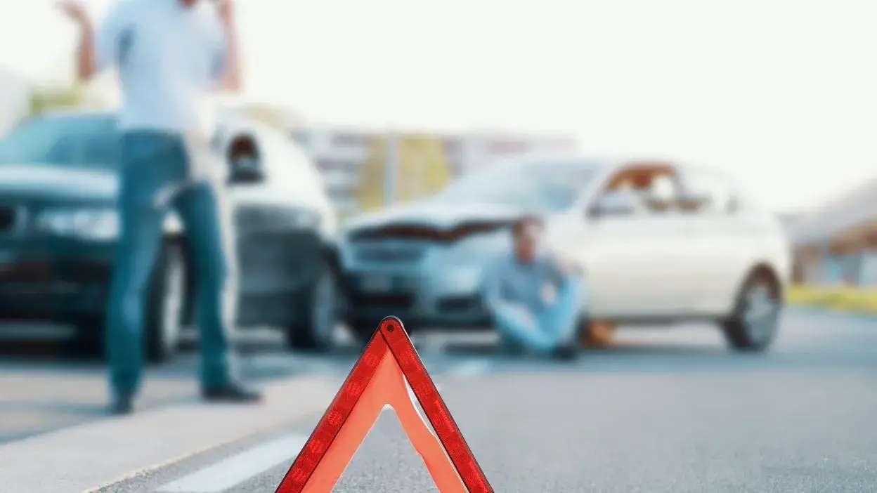 Triângulo de sinalização refletor colocado na estrada com dois carros envolvidos em acidente e pessoas ao fundo