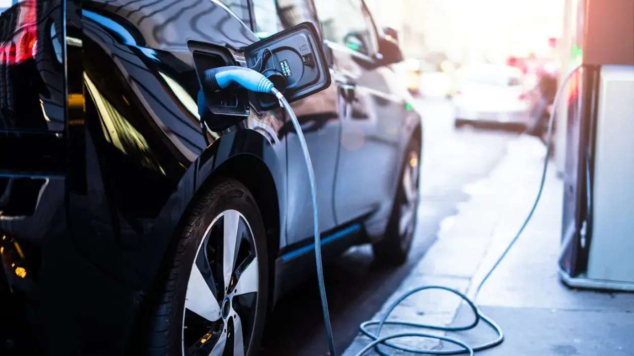 Carro elétrico preto ligado a posto de carregamento numa rua urbana.