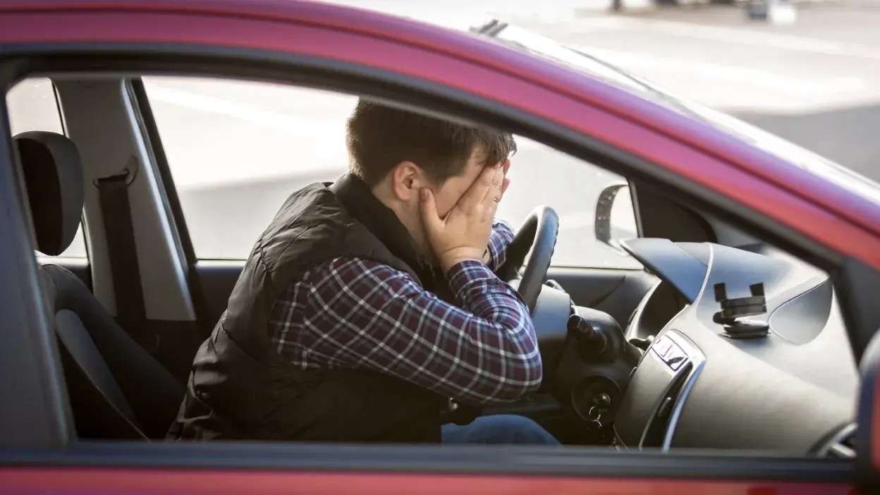 Homem sentado no banco do condutor com as mãos no rosto, aparentando stress ou ansiedade dentro de um carro vermelho.