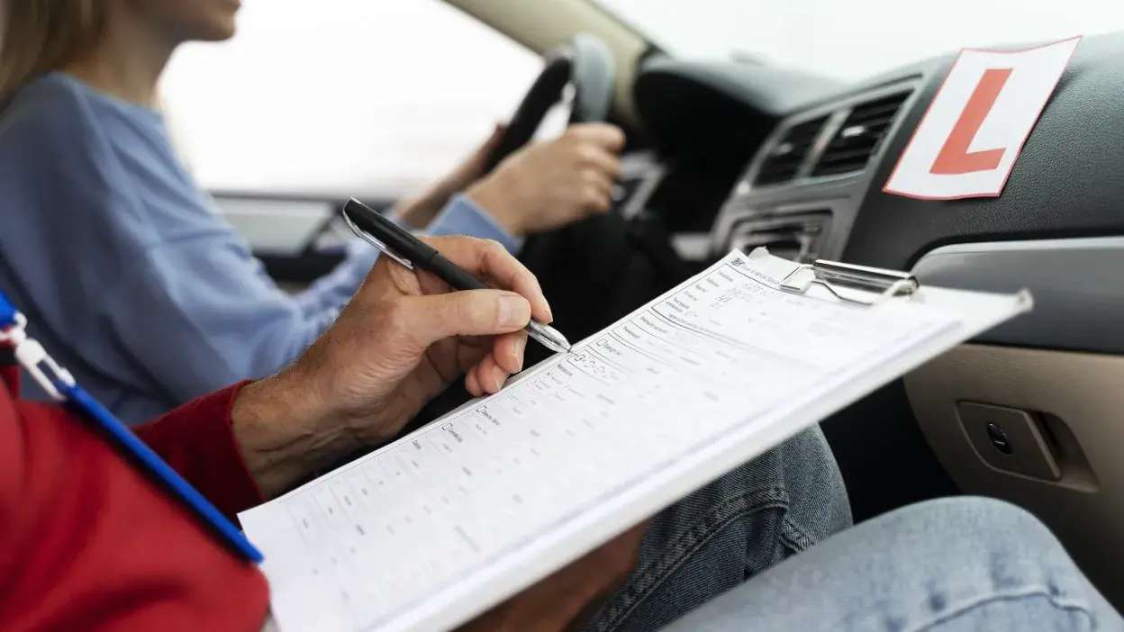 Instrutor a preencher ficha durante exame de condução com condutor ao volante e placa de aprendiz no carro