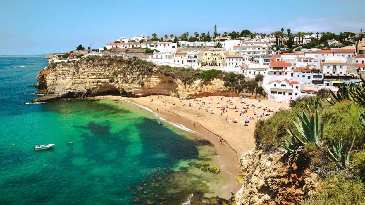 Praia com areia dourada, falésia rochosa e casas brancas junto ao mar com água verde-turquesa.