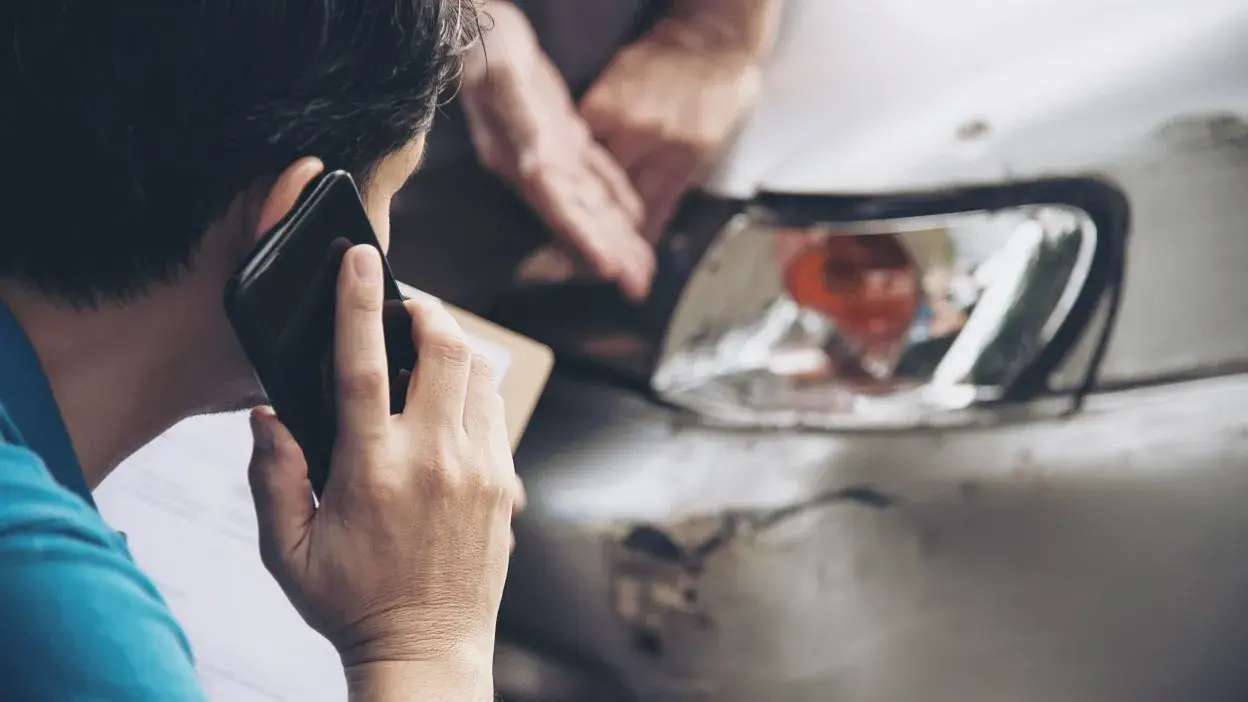 Pessoa ao telefone junto a carro com danos na frente após acidente