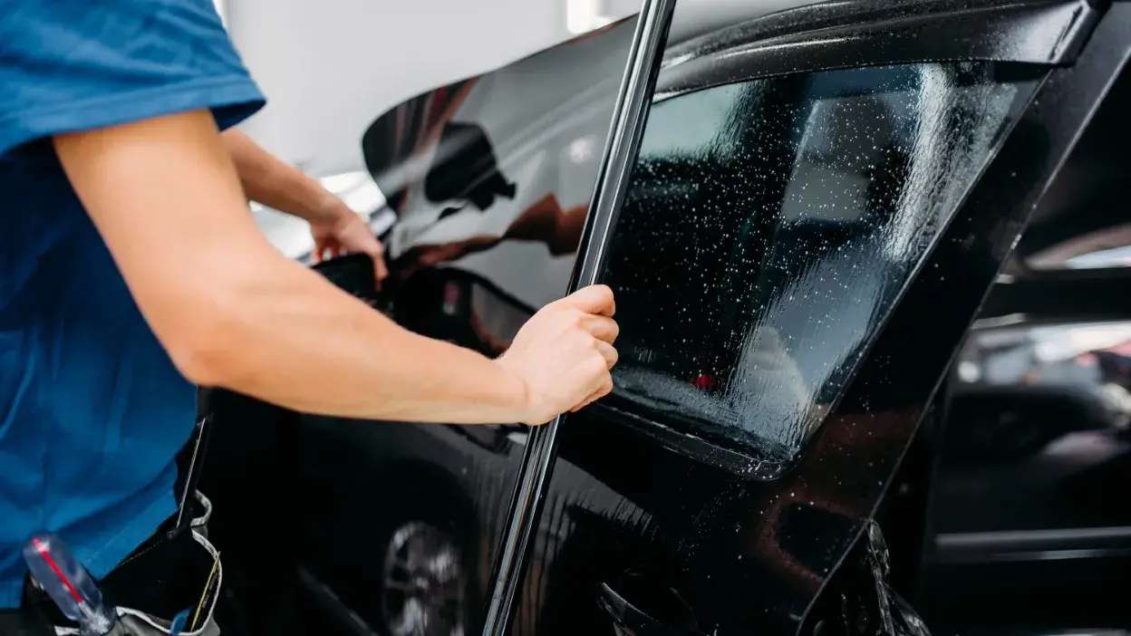 Pessoa a aplicar película numa janela de carro preto com gotas de água na superfície.