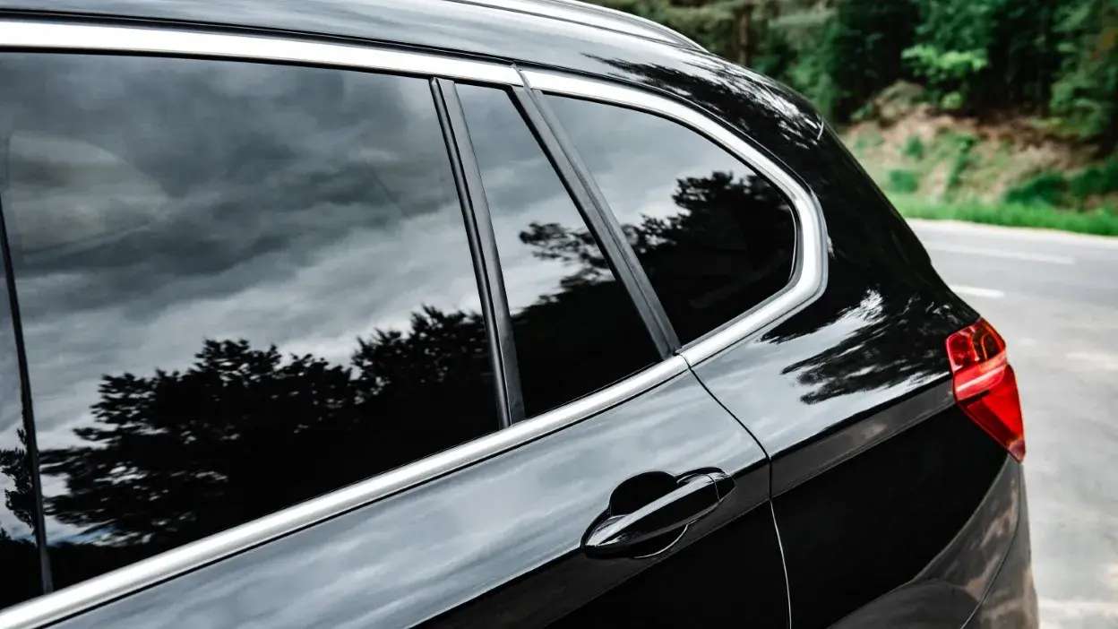 Vidros traseiros de um carro preto com reflexo de árvores e céu nublado.