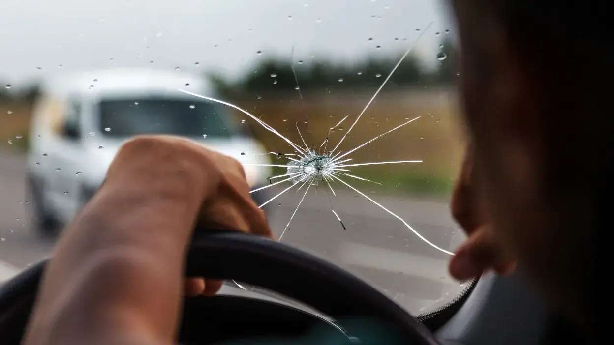 Vidro dianteiro de carro partido com mão no volante e veículo ao fundo desfocado