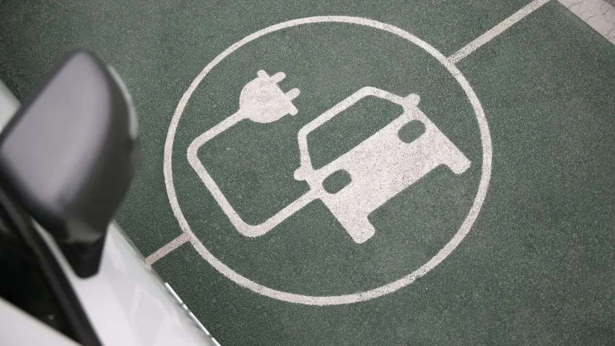 Sinalização no chão para lugar de carregamento de carros elétricos com símbolo de carro e ficha elétrica branca sobre pavimento verde.