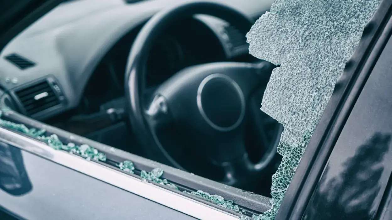 Vidro partido da janela lateral de um carro com estilhaços no interior e volante visível.