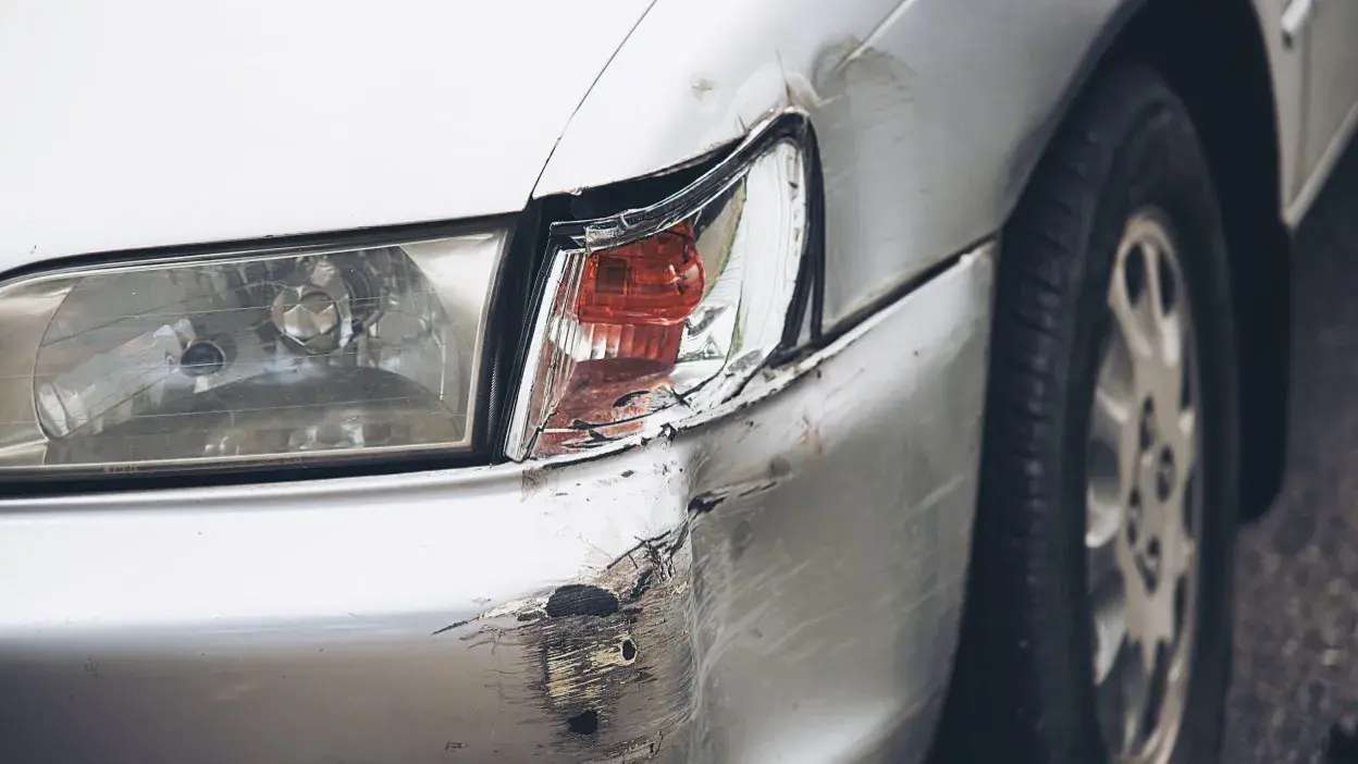 Dano visível no para-choques e farolim traseiro de um carro branco, com riscos e amolgadelas.