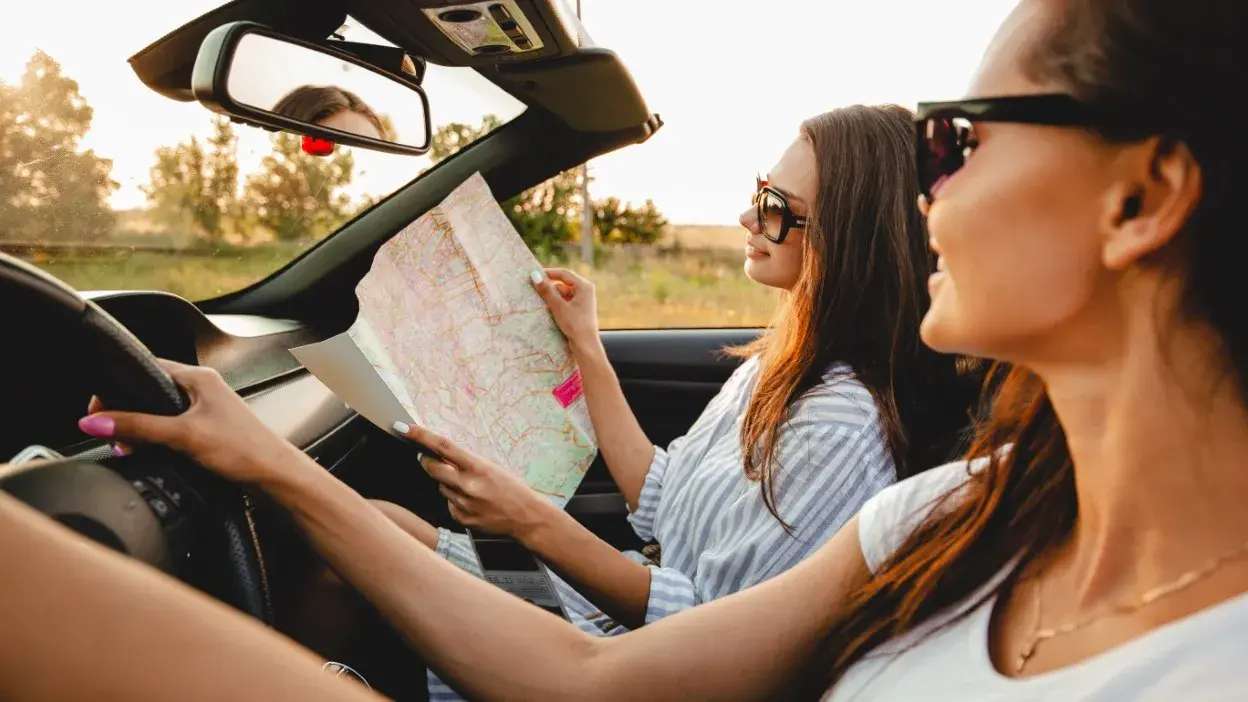 Duas mulheres de óculos de sol no carro, uma conduz e a outra lê um mapa.