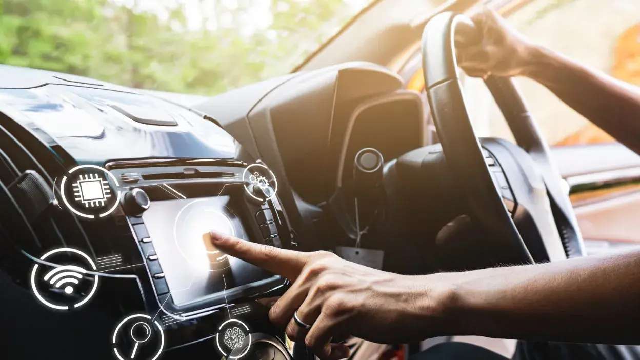 Mão a tocar no ecrã do computador de bordo de um carro com ícones digitais sobrepostos.