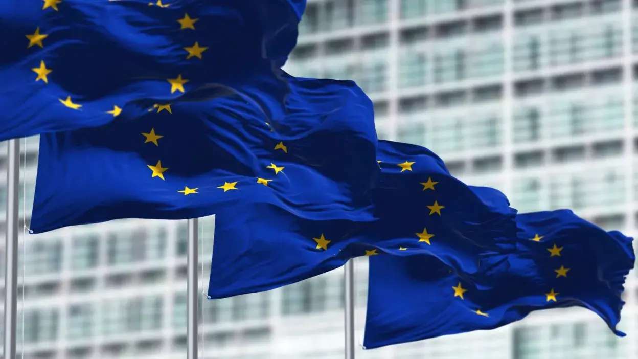 Bandeiras da União Europeia a ondular em frente a um edifício moderno com janelas de vidro.