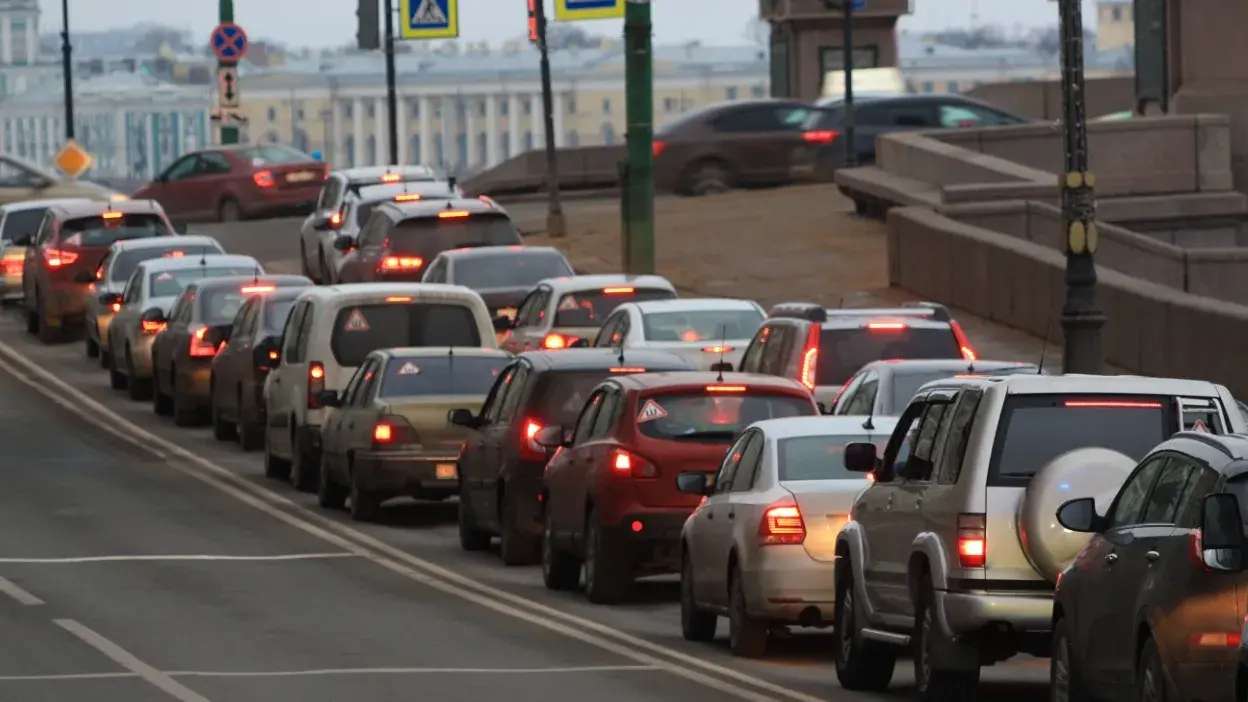 Vários carros parados em fila numa estrada urbana com sinais de trânsito visíveis ao fundo.