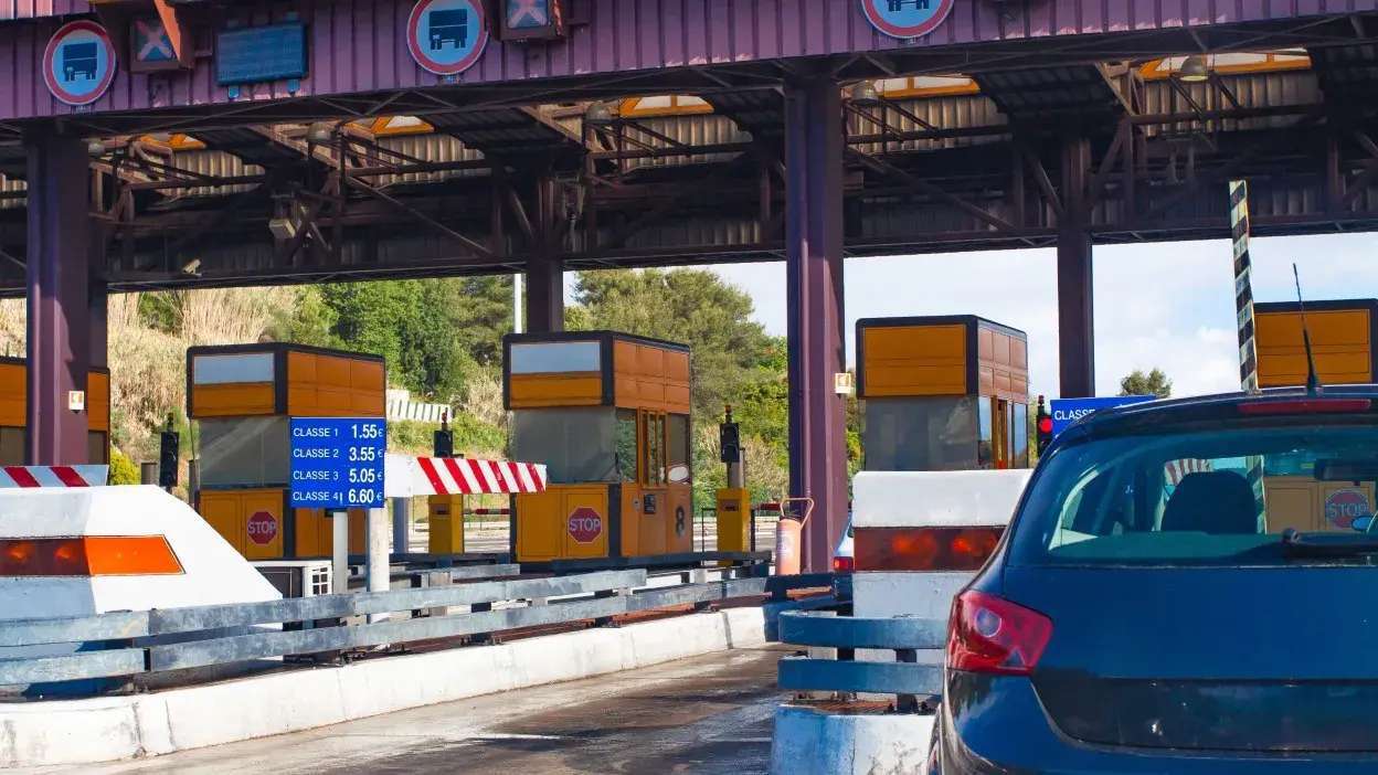 Cabines de portagem com sinais de paragem e tabela de preços para diferentes classes de veículos, com um carro azul à espera.