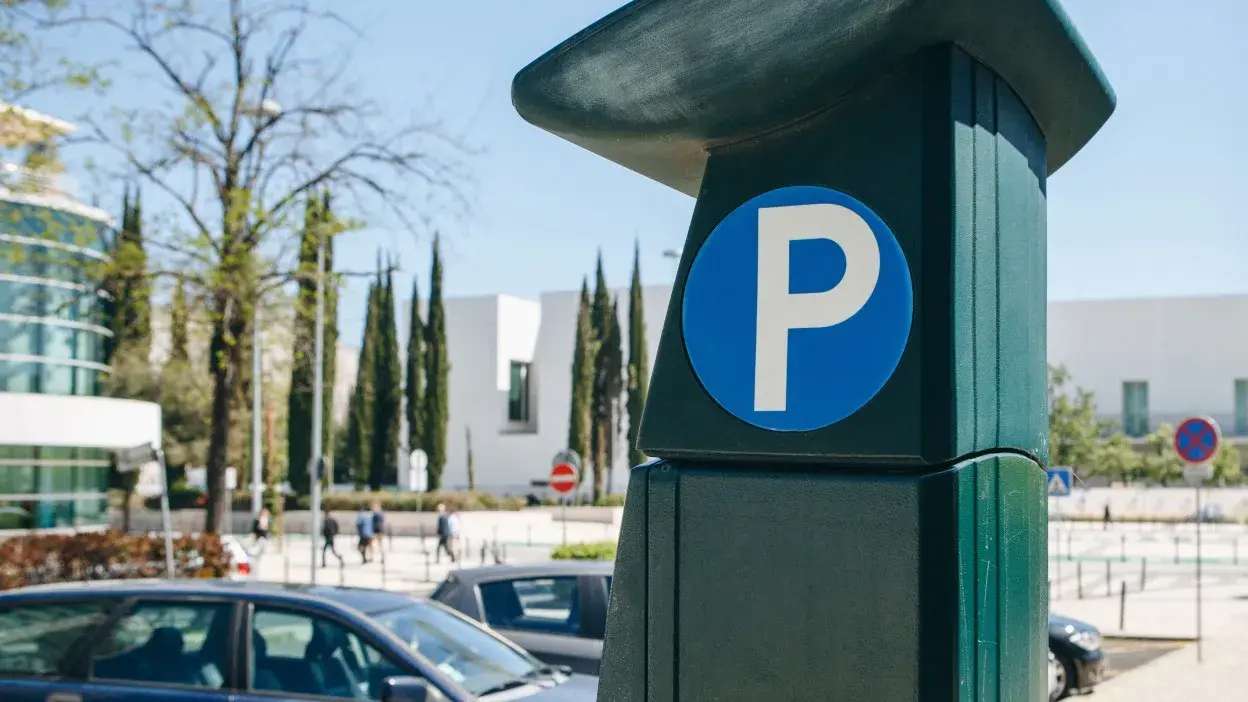 Parquímetro com símbolo de estacionamento azul numa rua com carros estacionados e edifícios ao fundo