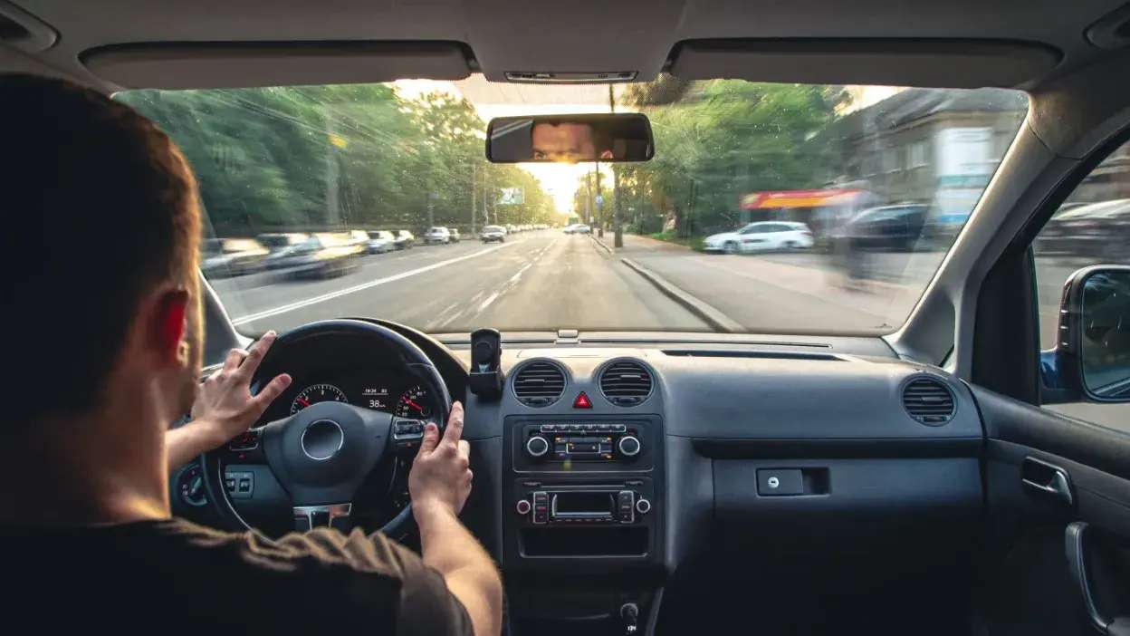 Homem a conduzir carro numa estrada urbana com trânsito e árvores ao fundo