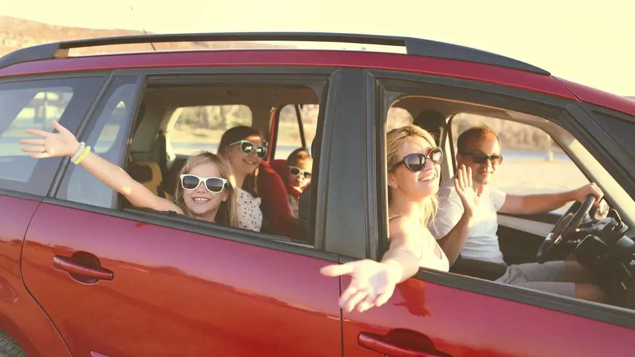Família sorridente com óculos de sol dentro de carro vermelho a acenar pela janela aberta.