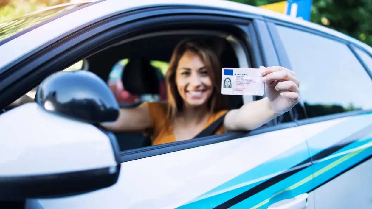 Mulher sorridente dentro de carro a mostrar carta de condução pela janela aberta.